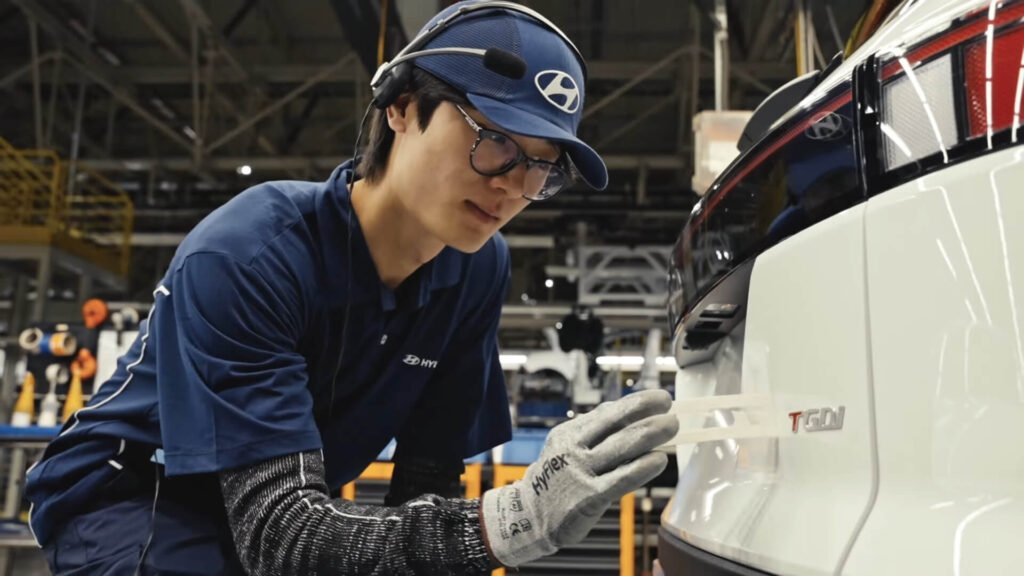 Homem remove adesivo que envolve emblema 'TGDI' de carro em linha de produção da Hyundai durante visitas guiadas na fábrica.